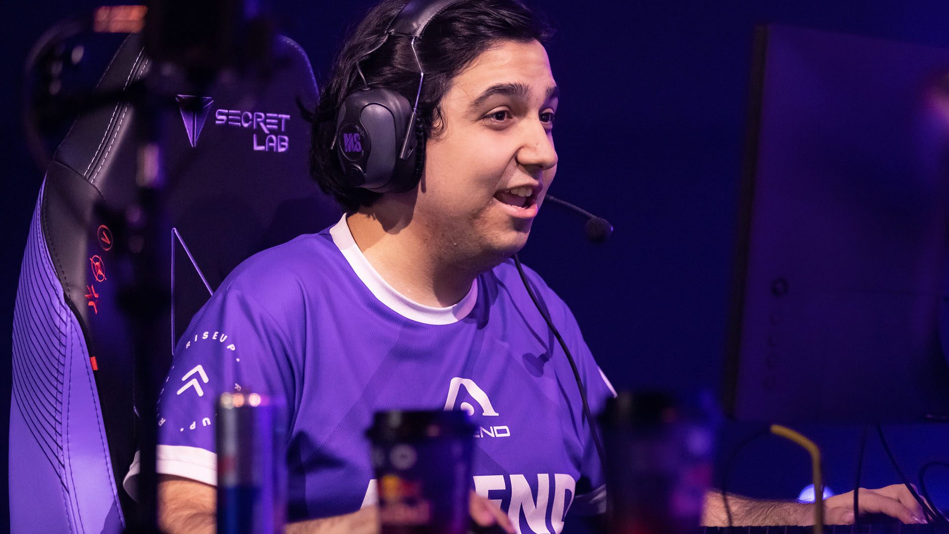 A player is enjoying the game and is sitting on a chair. He has mic headphones in his ears and a monitor in front of him. There are 2 coffee cups and one energy drink on the table