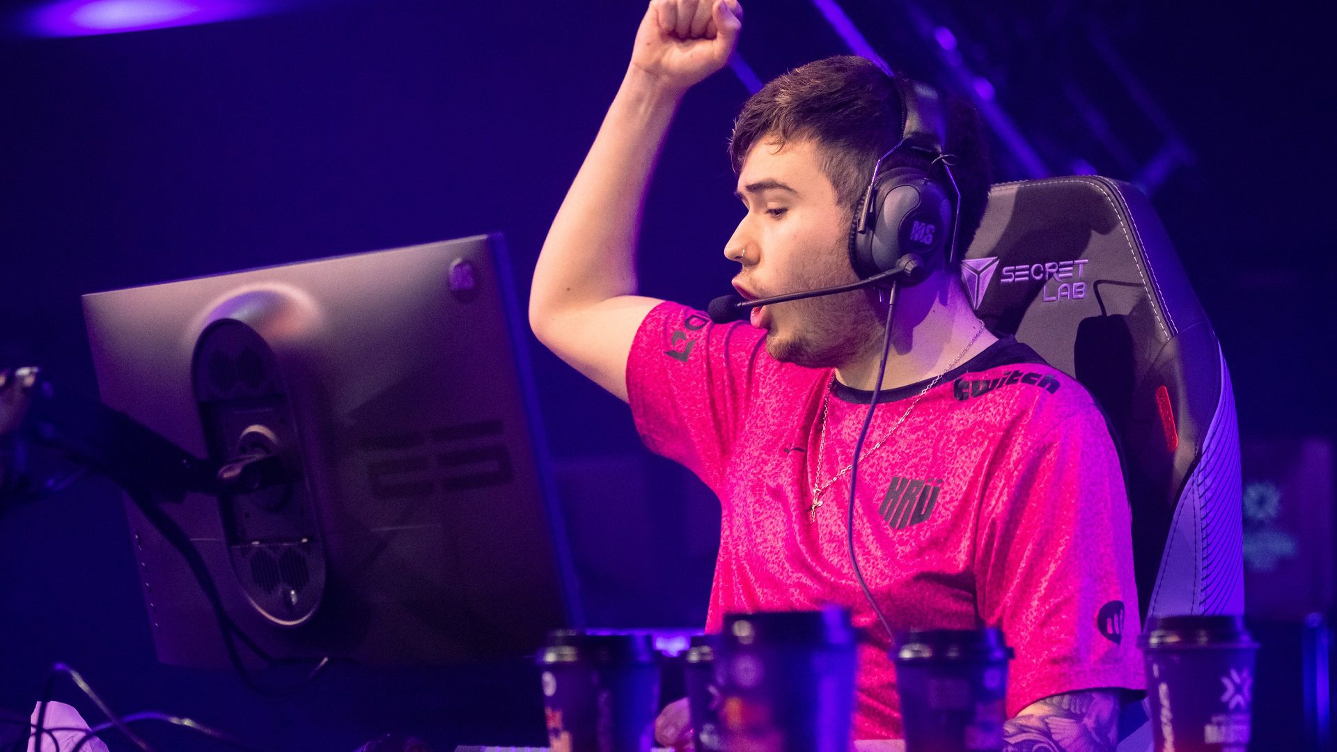 A valorant player looks very happy, and he has raised his hand with his one handful. He is sitting on a chair. He has mic headphones in his ears and a monitor in front of him. There are five coffee cups on the table
